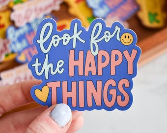 Puede incluir: Una pegatina de vinilo troquelada con fondo azul que presenta el texto "look for the HAPPY THINGS" en una mezcla de letra cursiva azul claro y letras de bloque rosas, acompañada de una carita sonriente amarilla y un icono de corazón amarillo.