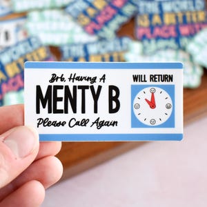 May include: A rectangular sticker with a white background and blue borders. It features the black text 'Brb, Having A MENTY B Please Call Again' and 'WILL RETURN' above a clock graphic with a red hand and small smiley face icons.