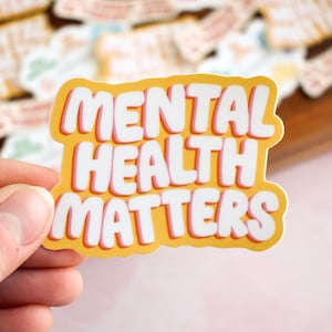 May include: A die-cut sticker featuring the text MENTAL HEALTH MATTERS in bold, white, rounded capital letters. The text has a soft pink outline and is set against a solid, mustard yellow background with a wavy, organic border shape.