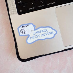 May include: A die-cut sticker featuring a white milk carton with the text MILK, 1%, and BETTER EVERY DAY, with a blue-outlined puddle shape containing the text EMBRACE MESSY ACTION, applied to the silver surface of a laptop keyboard area.