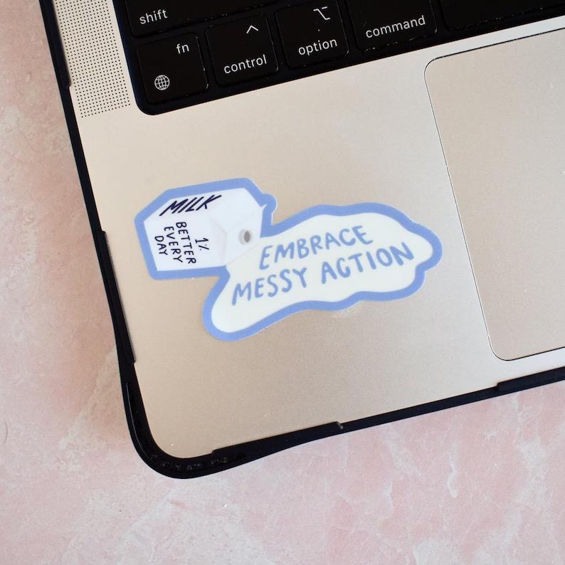 May include: A die-cut sticker featuring a white milk carton with the text MILK, 1%, and BETTER EVERY DAY, with a blue-outlined puddle shape containing the text EMBRACE MESSY ACTION, applied to the silver surface of a laptop keyboard area.