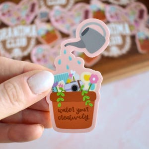 May include: A sticker featuring a brown flower pot with the text "water your creativity." A gray watering can pours water onto the pot, which contains a camera, flowers, and art supplies. The sticker is pink and has a light pink border.