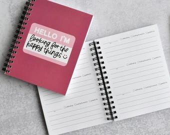 May include: A dark pink spiral-bound notebook with a white name tag graphic on the cover that reads "HELLO I'M looking for the happy things" with a smiley face. The open notebook shows lined pages with checkboxes for celebrating, looking forward to, and grateful for.