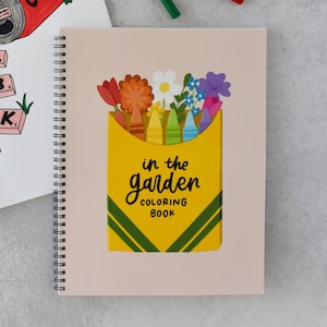 May include: A spiral-bound coloring book with a light beige cover featuring a yellow crayon box illustration. The box contains colorful crayons and various flowers, with the black text in the garden COLORING BOOK printed on the front.