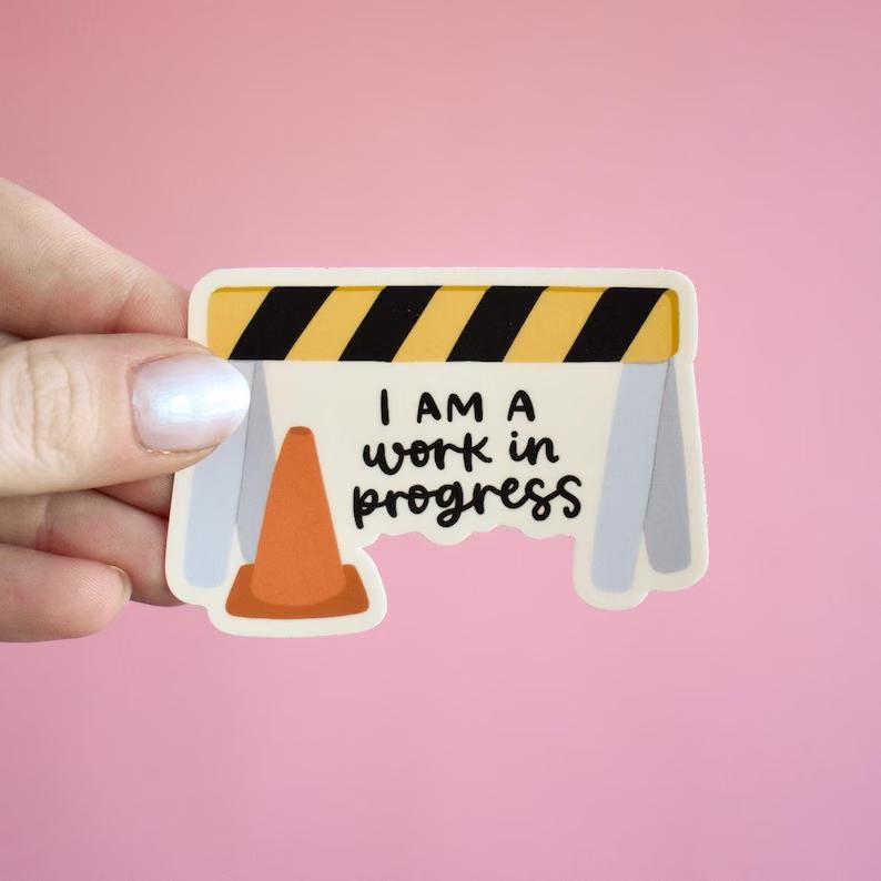 May include: A die-cut sticker featuring a construction barricade with yellow and black diagonal stripes, a light gray frame, and an orange traffic cone. The white center of the barricade displays the black text I AM A work in progress.