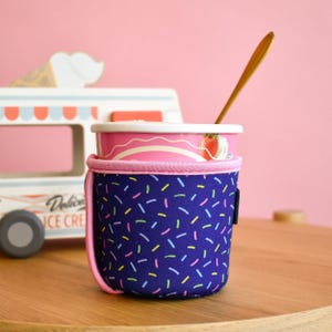 May include: A navy blue insulated cup holder with a colourful sprinkle pattern, holding a pink ice cream cup with a strawberry design. A gold spoon rests inside the cup. The background includes a toy ice cream van.