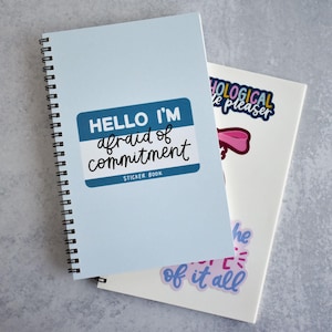 May include: A blue spiral bound notebook with a sticker that says "Hello I'm afraid of commitment" and a sticker book label. The notebook is on top of a white notebook with stickers that say "hological le pleaser" and "he of it all".