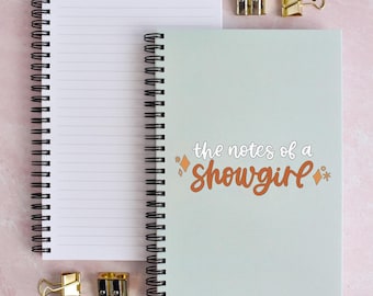 May include: A spiral-bound notebook with a light green cover featuring the text "the notes of a showgirl" in white and orange script, accompanied by small orange star and diamond icons, alongside a second open notebook and two metal sharpeners.