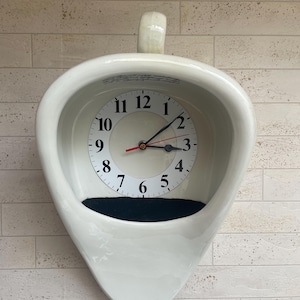 Könnte beinhalten: Eine weiße Keramikuhr in Form eines Urinals, an einer hellen Backsteinmauer befestigt. Das Zifferblatt hat schwarze Ziffern und Zeiger, mit einem schwarzen Bereich am Boden. Eine skurrile, neuartige Uhr.