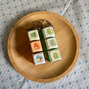 May include: Two keychains with colorful keycaps. One keychain has three keycaps with a frog and other cartoon characters. The other keychain has three keycaps with a frog and other cartoon characters. The keychains are on a wooden plate.
