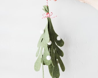 May include: A felt mistletoe ornament featuring multiple olive green leaves with rounded edges and small white felt berries attached. The ornament is tied at the top with a red and white baker's twine string, forming a small bow for hanging.