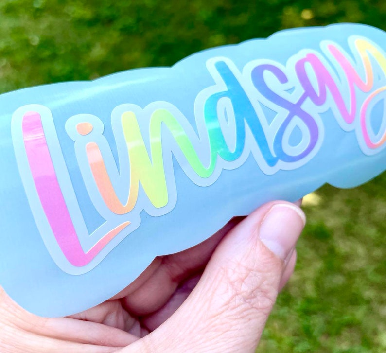May include: A light blue water bottle with the name "Lindsay" in a rainbow holographic font. The text is in a cursive style, with a white outline. The bottle is held against a blurred green background.