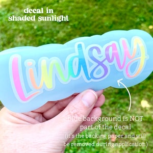 May include: A light blue decal with the name "Lindsay" in a rainbow gradient font. The text "decal in shaded sunlight" is above the name. The text "blue background is NOT part of the decal" is below the name.