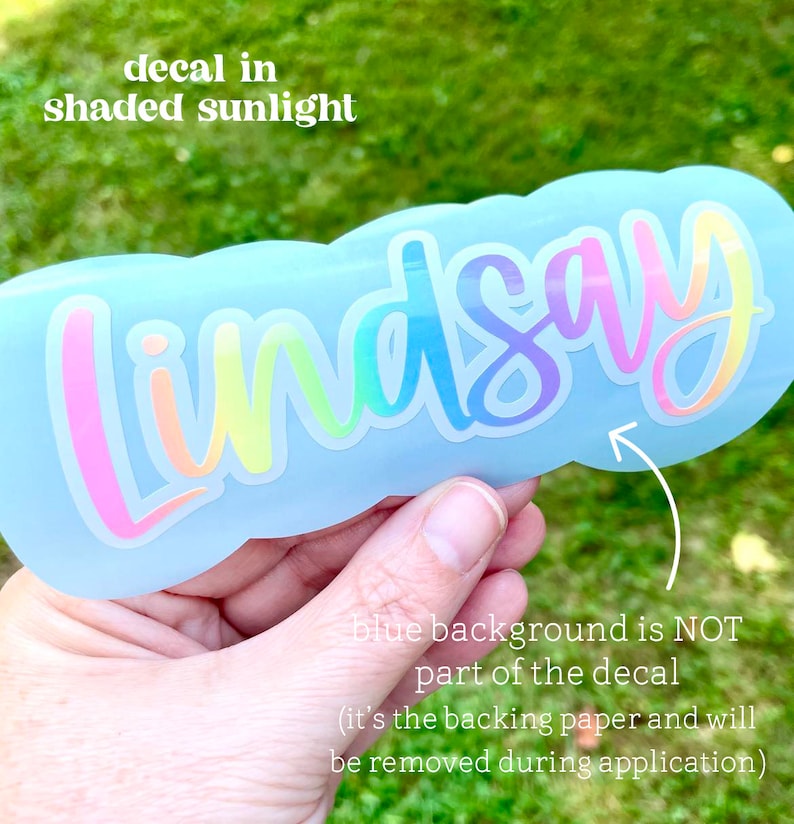 May include: A light blue decal with the name "Lindsay" in a rainbow gradient font. The text "decal in shaded sunlight" is above the name. The text "blue background is NOT part of the decal" is below the name.