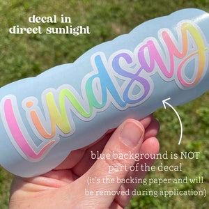 May include: A decal with the name "Lindsay" in a rainbow gradient font. The decal is shown on a light blue surface. Text in the image reads "decal in direct sunlight" and "blue background is NOT part of the decal (it's the backing paper and will be removed during application)".