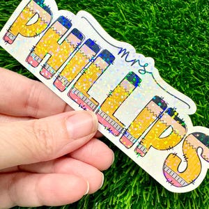 May include: A holographic sticker featuring the name PHILLIPS in large, yellow pencil-shaped letters with black tips and pink erasers. The word mrs is written in a cursive, dark blue script above the letters. The entire sticker has a glittery finish.