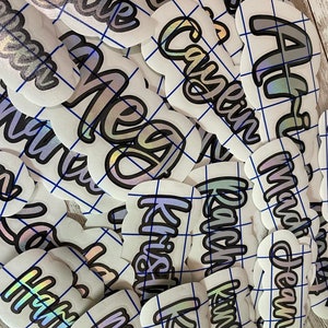 May include: A collection of white, die-cut stickers with names in a cursive font. The names are outlined in black with a silver and iridescent fill. The stickers are arranged on a blue grid.