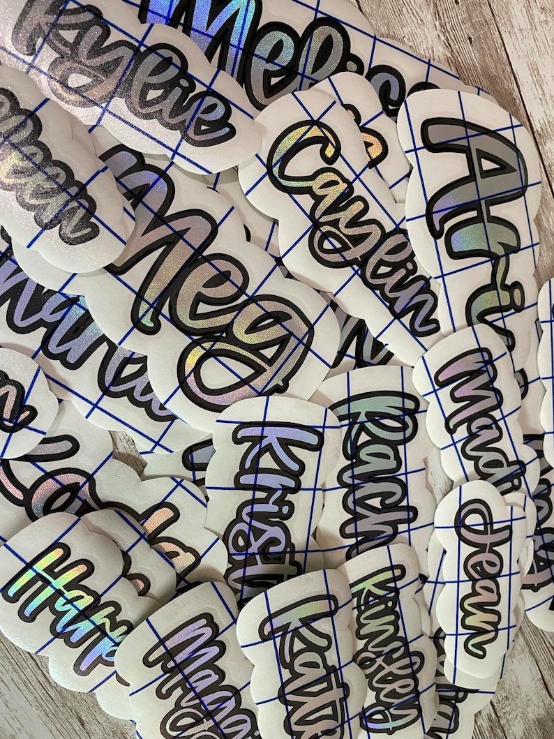 May include: A collection of white, die-cut stickers with names in a cursive font. The names are outlined in black with a silver and iridescent fill. The stickers are arranged on a blue grid.