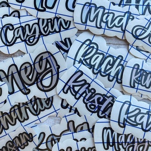 May include: A collection of white, cloud-shaped vinyl stickers with various names in a cursive font. The names are in a gradient color scheme with a black outline. Each sticker has a blue grid pattern.