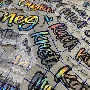 May include: A collection of iridescent name decals with a holographic effect. The names are written in a stylized font with a black outline. The decals are on a white background with a blue grid pattern.