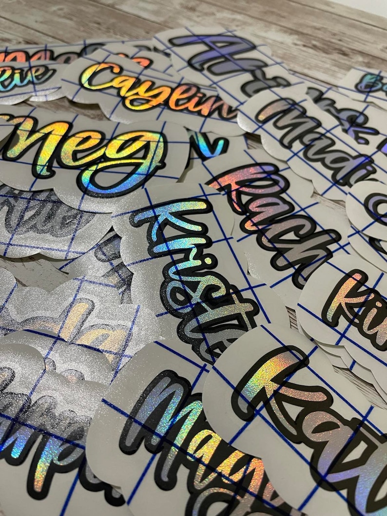 May include: A collection of iridescent name decals with a holographic effect. The names are written in a stylized font with a black outline. The decals are on a white background with a blue grid pattern.