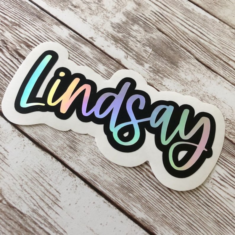 May include: A white sticker with the name "Lindsay" in a holographic, rainbow-colored script font, outlined in black. The sticker has a unique, curved shape and is placed on a light-colored, wooden surface.