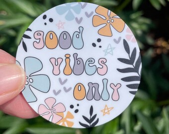 May include: A circular sticker with a white background featuring the text "good vibes only" in a multi-colored, rounded font. The design includes various illustrations of flowers, leaves, small stars, and dots in shades of pink, blue, orange, and gray.