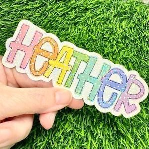 May include: A die-cut sticker featuring the name HEATHER in a bold, sans-serif font. Each letter displays a different pastel color, including pink, orange, yellow, green, blue, and purple, all finished with a shimmering, holographic glitter effect.