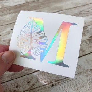 May include: A white sticker with the letter "M" in a holographic, iridescent finish. A detailed illustration of a monstera leaf partially overlays the letter. The sticker is held against a light wood surface.
