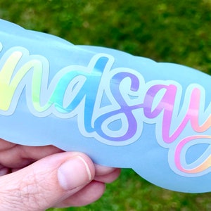 May include: A light blue sticker with the name "Lindsay" in a rainbow holographic font. The sticker has a cloud-like shape and is held against a blurred green background.