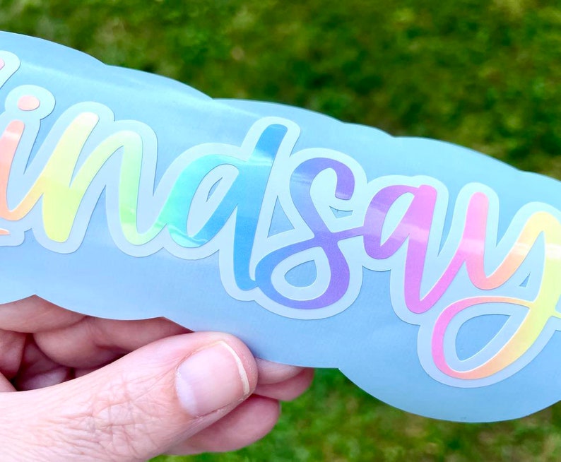 May include: A light blue sticker with the name "Lindsay" in a rainbow holographic font. The sticker has a cloud-like shape and is held against a blurred green background.