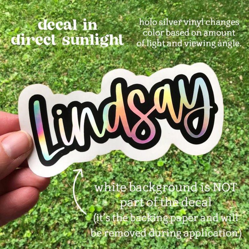 May include: A holographic vinyl decal with the name "Lindsay" in a script font, outlined in black and white. The decal's color shifts based on light and viewing angle. Text on the decal states it changes color in direct sunlight.
