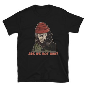 Op de afbeelding: Zwart T-shirt met een vintage illustratie van een chimpansee die een rode hoed draagt. De tekst "Are We Not Men?" is onder de afbeelding gedrukt.