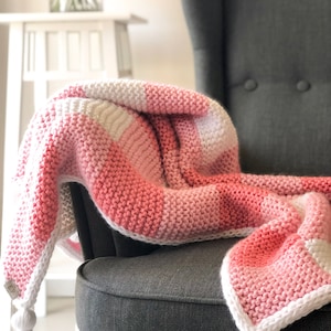 May include: A pink and white crocheted throw blanket with white tassels on a grey armchair.