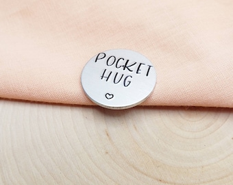 May include: A silver metal disc with the words "Pocket Hug" engraved on it. A small heart symbol is also engraved on the disc.