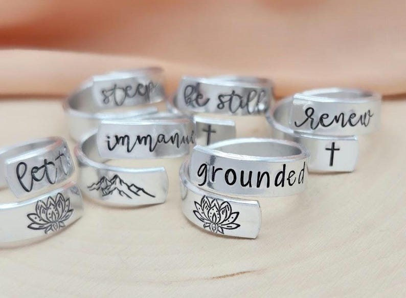 May include: A set of silver rings with different inspirational words and designs engraved on them. The words include "let it be", "immanuel", "be still", "renew", "deep", and "grounded". Some rings feature a mountain range, a cross, and a lotus flower.