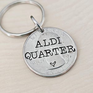 May include: A silver key chain with a quarter attached to it. The quarter has the words "ALDI QUARTER" engraved on it with a small heart.
