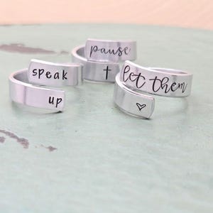May include: Three silver rings with inspirational messages engraved on them. The rings read "Speak Up", "Pause", and "Let Them".