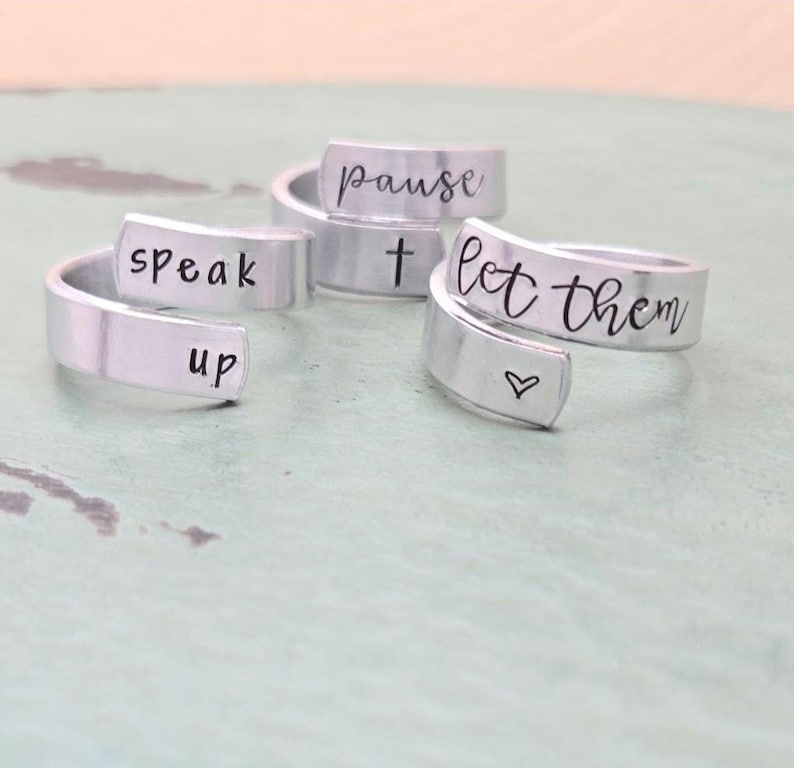 May include: Three silver rings with inspirational messages engraved on them. The rings read "Speak Up", "Pause", and "Let Them".