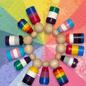 May include: A circle of wooden figures painted with various pride flags, including the rainbow, transgender, bisexual, and asexual flags.
