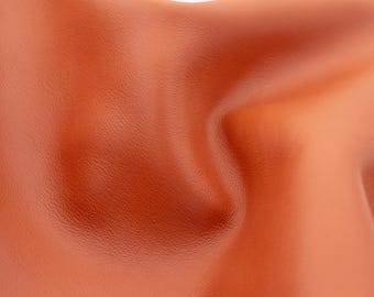 Może przedstawiać: Zbliżenie na gładki, elastyczny arkusz pomarańczowo-brązowej skóry. Materiał ma naturalną, drobnoziarnistą fakturę z miękkimi fałdami i krzywiznami, które tworzą subtelne refleksy i cienie na całej powierzchni tego materiału.