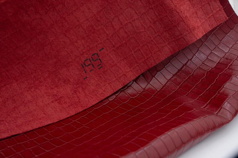 May include: A close-up of two pieces of red fabric. The top piece is a soft, textured fabric with the number "199" printed on it. The bottom piece is a smooth, embossed fabric with a crocodile pattern.