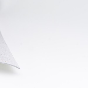 Pode incluir: Um close-up de um peda&ccedil;o de material cinza claro e texturizado com uma borda curva. O material parece macio e ligeiramente salpicado, em um fundo branco brilhante.