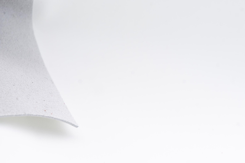 Pode incluir: Um close-up de um peda&ccedil;o de material cinza claro e texturizado com uma borda curva. O material parece macio e ligeiramente salpicado, em um fundo branco brilhante.