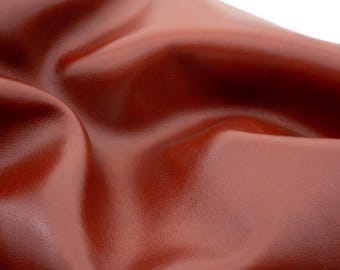 Może przedstawiać: Zbliżenie na gładki, bogaty czerwonobrązowy materiał skórzany z głębokimi, płynnymi fałdami i miękkimi krzywiznami. Powierzchnia ma subtelny połysk, który podkreśla naturalną teksturę i spójny kolor materiału na całej jego falistej powierzchni.