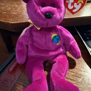Può includere: Orso di peluche viola Beanie Baby con un nastro dorato intorno al collo. L'orso ha un naso nero e occhi neri. L'orso ha un'etichetta che dice "Beanie Baby" e "Ty". L'orso ha una toppa sul petto che dice "Terra" e ha un'immagine della Terra con un sole che sorge alle sue spalle.