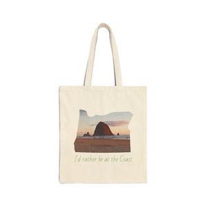 May include: Cream-colored tote bag featuring an Oregon outline with a coastal scene. The image shows a large rock formation on a beach at sunset. The text "I'd rather be at the Coast" is printed below the image.