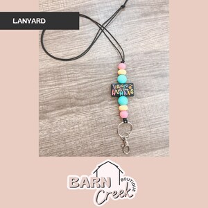 May include: Black lanyard with colorful beads and a rectangular charm that reads "Teacher of Tiny Humans." The lanyard has a metal clasp and key ring. The background is a light wood grain.