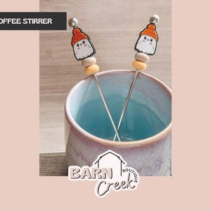 May include: Two coffee stirrers with a ghost design, each featuring a silver stem and a decorative top with a ghost wearing an orange hat. The stirrers are in a blue and purple mug. The text "COFFEE STIRRER" is in the top left corner.