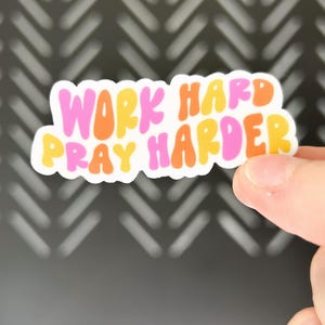 May include: A colorful sticker with the words "WORK HARD PRAY HARDER" in pink, orange, and yellow bubble letters. The sticker has a white border and is held against a dark background.